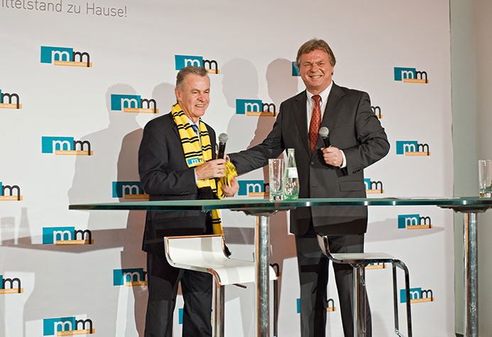 Fußballtrainer Ottmar Hitzfeld und m+m Geschäftsführer Michael Müller in einem Podiumsgespräch vor einer m+m Logowand.