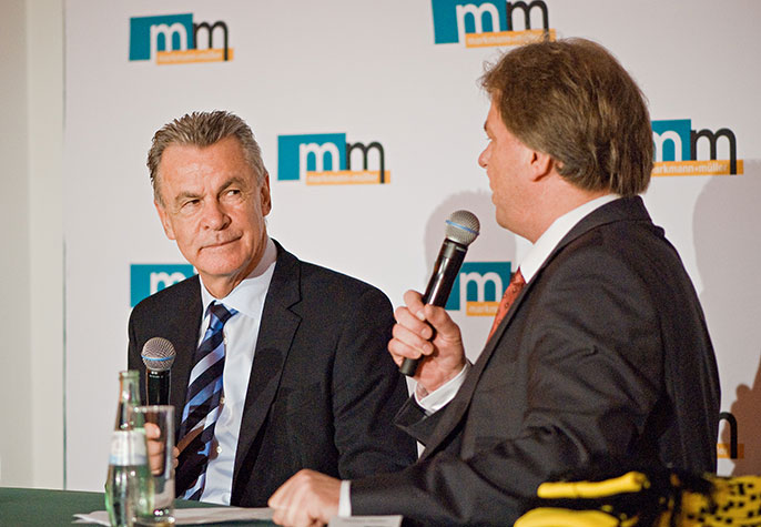 Fußballtrainer Ottmar Hitzfeld und m+m Geschäftsführer Michael Müller in einem Podiumsgespräch vor einer m+m Logowand.
